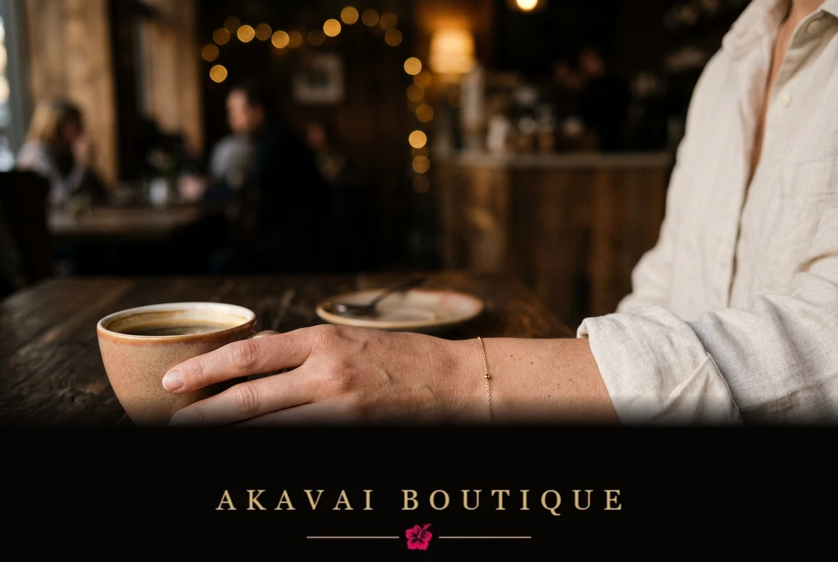 Bracelet fin doré porté au poignet, main près d’une tasse de café sur une table en bois, lumières chaudes en arrière-plan.