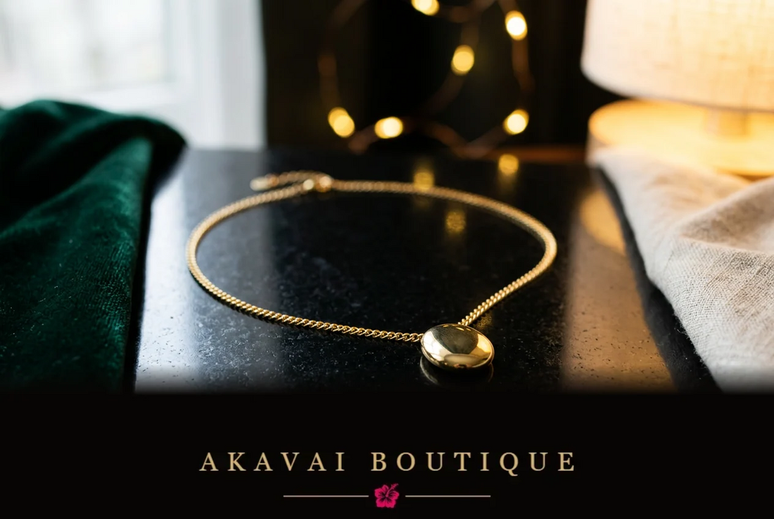 Collier doré à pendentif ovale sur une surface noire réfléchissante, entre un tissu vert émeraude et du lin écru, éclairé par une fenêtre et une lampe à la lumière chaude.