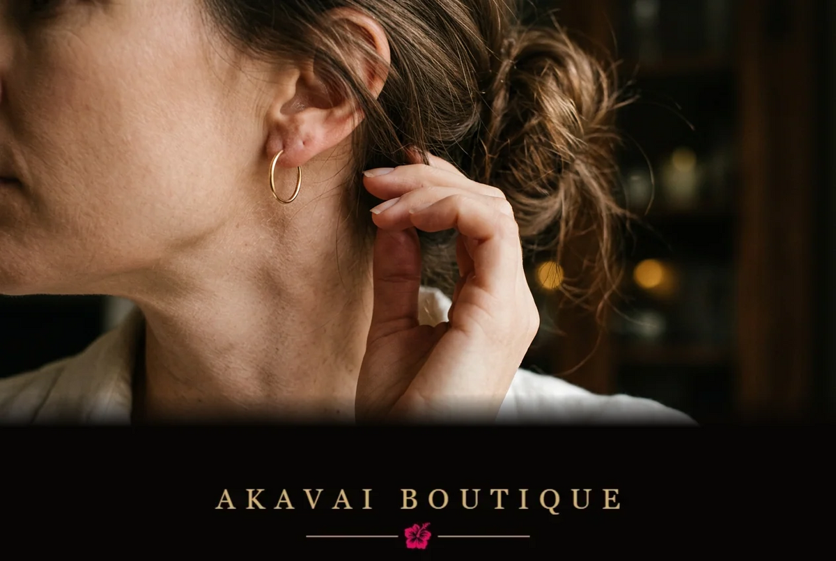 Créole fine dorée portée à l’oreille, cheveux relevés en chignon, main près du cou, chemise écrue et lumières chaudes en arrière-plan.